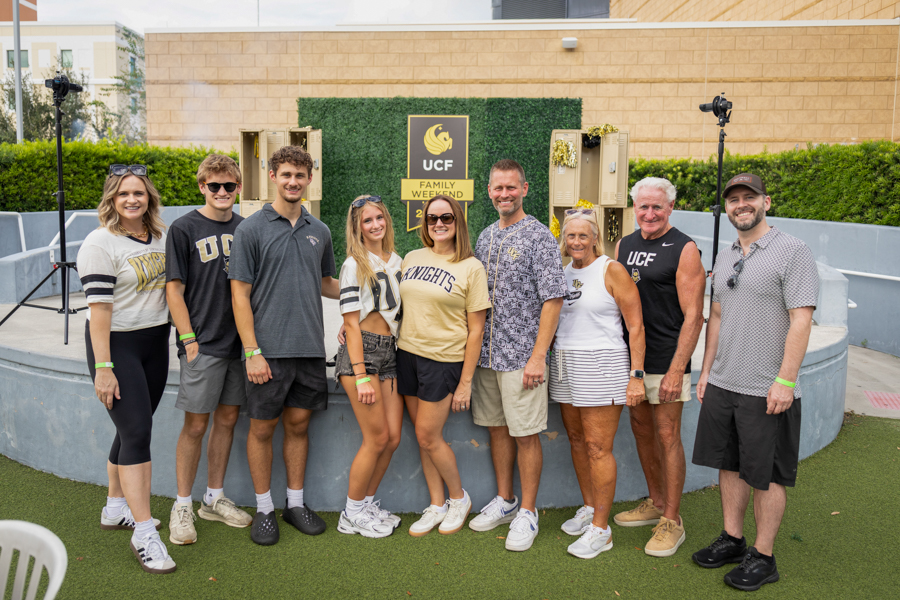 Group of eight adults and two younger people posing together outdoors in front of a UC Family Weekend sign at a campus venue, smiling and wearing casual athletic clothing.