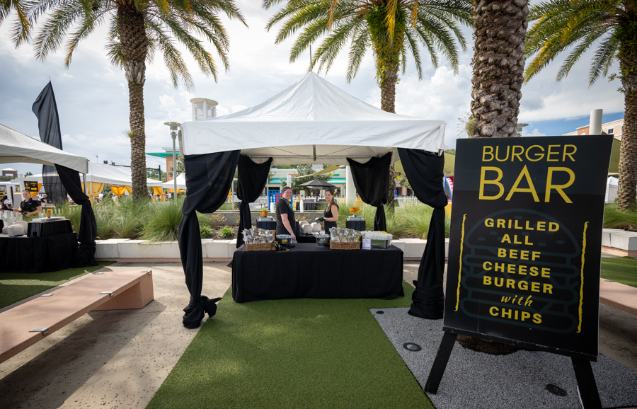 Outdoor burger stand under a white canopy at an event, with staff behind a black-draped counter and a tall burger bar sign nearby.