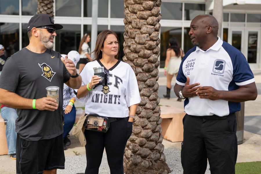 Three adults stand near palm trees, chatting at an outdoor event; two wear branded shirts and hold cups, while a concierge-like staff member in a polo talks nearby.