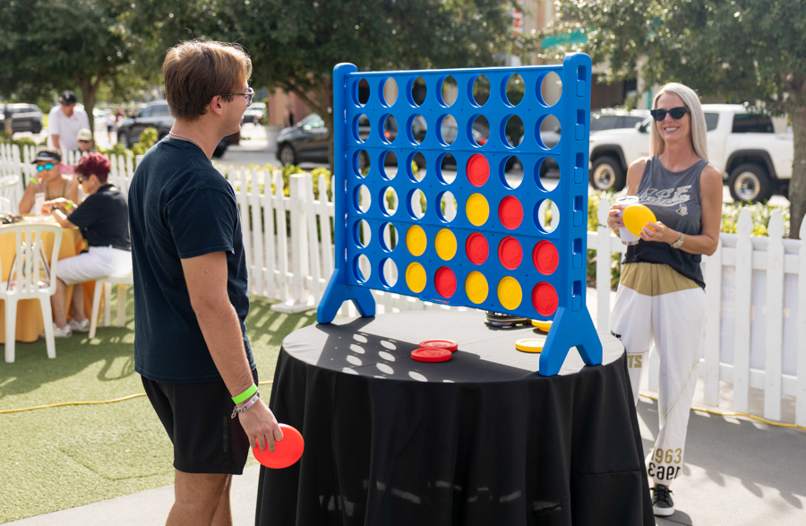 Two players stand at a life-size blue Connect Four game, holding red and yellow discs outdoors as others sit at tables nearby.