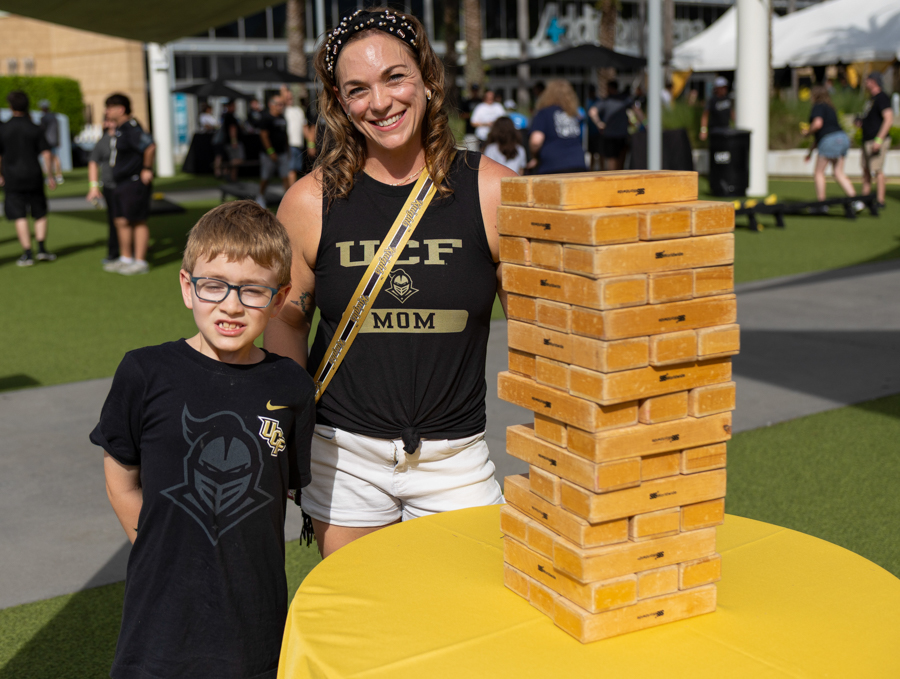 Smiling woman in a UCF MOM shirt and a boy pose beside a tall Jenga tower at an outdoor event.