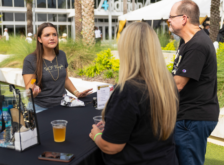 Woman at an outdoor booth hands a snack to two visitors across a black tablecloth. A cup of drink and a display with snacks are visible on the table.