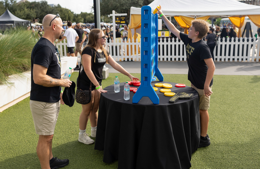 A group of three people gathered around a black-tableclothed round table at an outdoor event, taking turns throwing discs toward a tall blue vertical game tower.