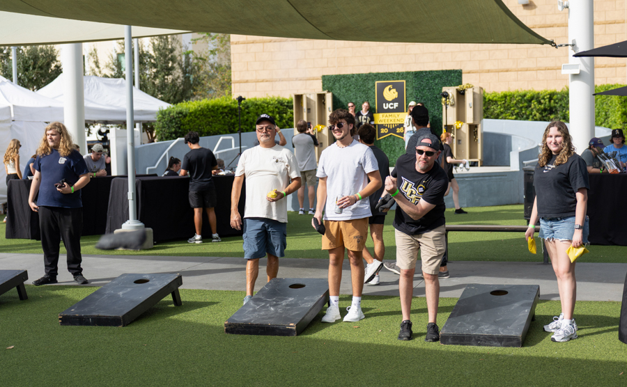 Group of people playing cornhole on a sunny outdoor lawn, tossing bags toward boards with a UC F banner in the background.