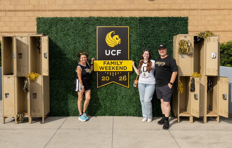 Four people pose in front of a UCF Family Weekend banner on a hedge wall, with gold lockers on either side.