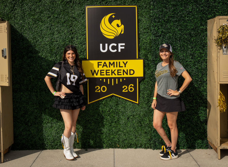 Two women pose beside a UCF Family Weekend 2026 banner on a green hedge wall, smiling at the camera.