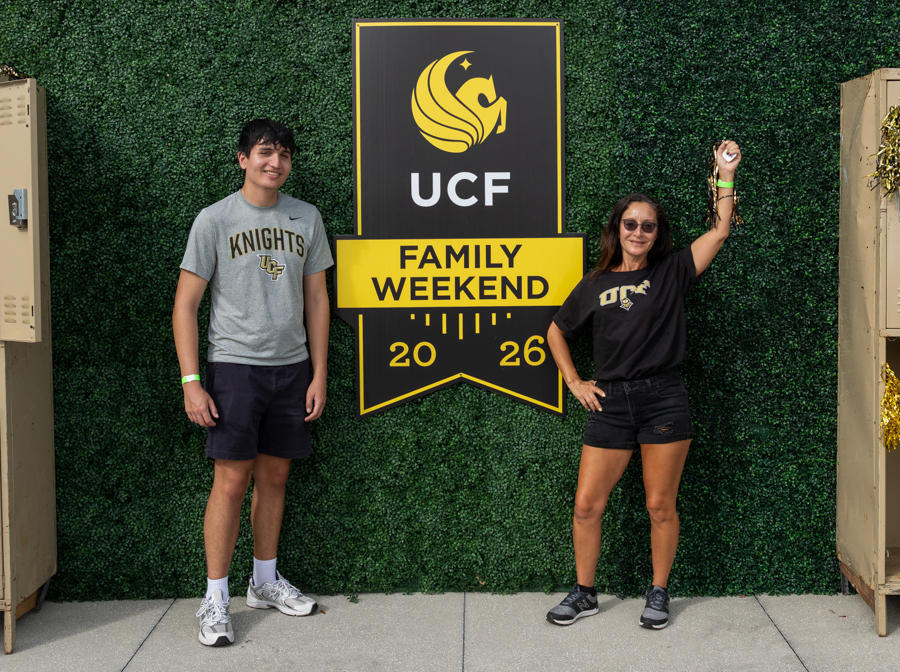 Two young adults stand on either side of a UCF Family Weekend sign against a green hedge backdrop, 2026 visible on the banner.