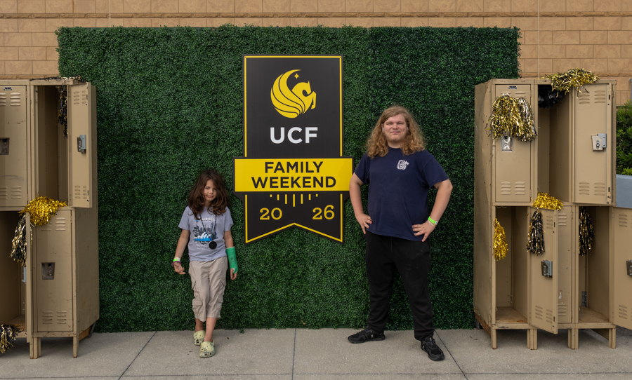 Two children stand in front of a UCF Family Weekend banner against a green hedge backdrop with lockers on either side.