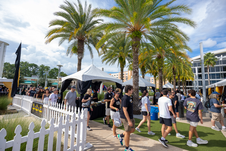 Outdoor festival or market with white tents, a white fence, and palm trees on a sunny day with visitors walking by.