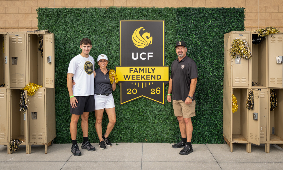Three people pose in front of a UCF Family Weekend 2026 banner on a green hedge backdrop, with lockers on both sides of the display.