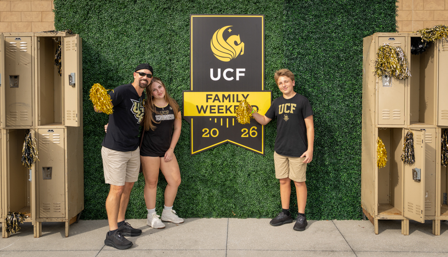 Family posing in front of a UCF Family Weekend banner with lockers on both sides; two adults and a teen in matching UCF shirts and khaki shorts hold gold pom-poms.