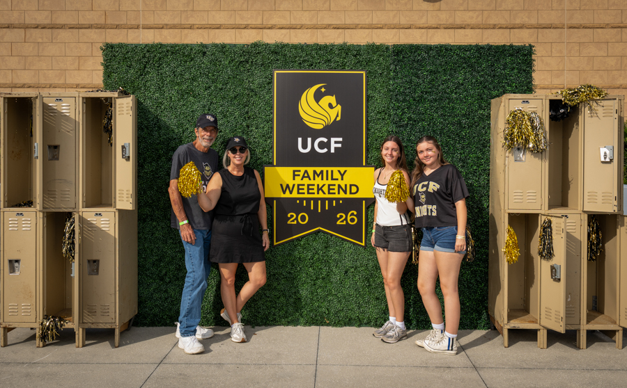 Five people pose in front of a UCF Family Weekend banner on a green hedge backdrop, with gold pom-poms and lockers on either side outside a building.