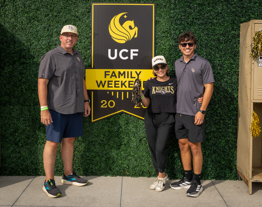 Three people pose and smile in front of a UCF 'Family Weekend' banner mounted on a green hedge wall, wearing Knights apparel and casual athletic shoes (2020).