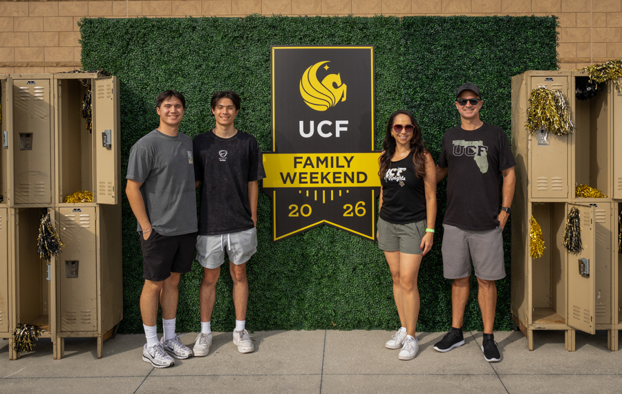 Four people pose in front of a UCF Family Weekend sign on a green hedge backdrop, with beige lockers on each side.