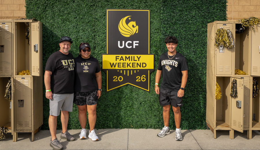 Three people wearing UCF gear pose in front of a green ivy backdrop and a UCF Family Weekend banner (2026).