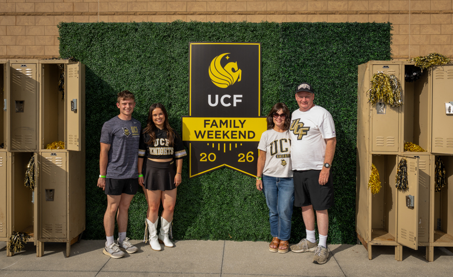 Family of four posing in front of a UCF Family Weekend sign on a green hedge backdrop, lockers on both sides, wearing UCF apparel and casual summer outfits.