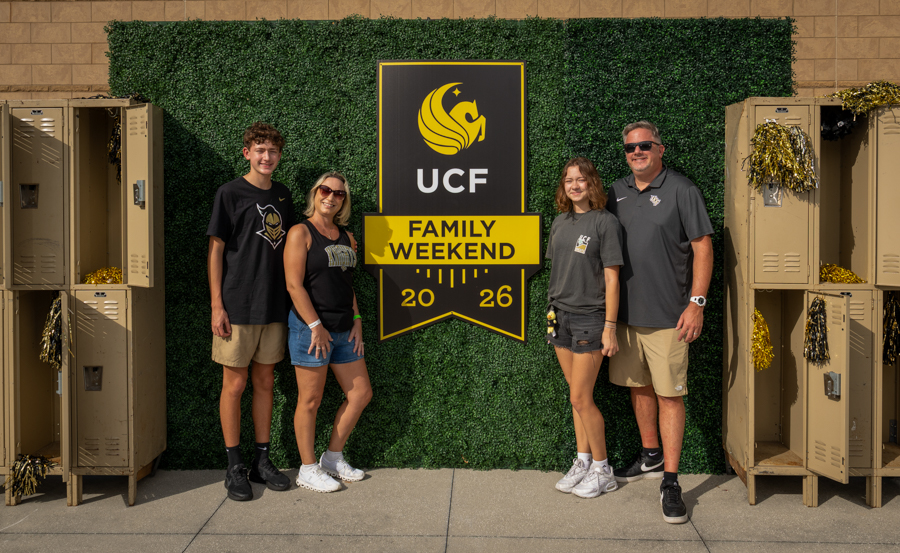 Family of four posing in front of a UCF Family Weekend 2026 sign with gold decorations and lockers on either side.