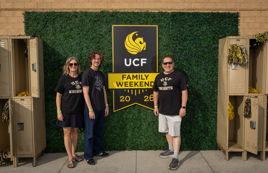 Three people stand in front of a UCF Family Weekend banner against a green hedge, wearing UCF Knights shirts and smiling.