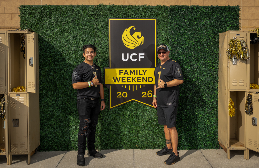Two men pose beside a UCF Family Weekend banner on a green hedge wall, with lockers and gold decorations in the scene.
