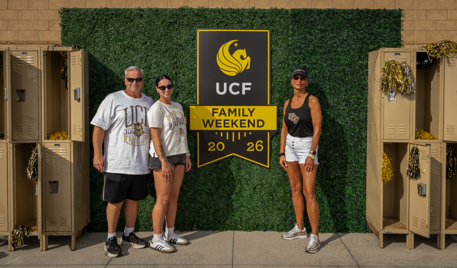Three people pose next to a UCF Family Weekend 2026 sign against a hedge backdrop, with beige lockers on both sides.
