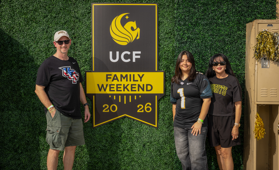 Three people pose beside a large UCF Family Weekend 2026 sign against a green hedge wall, outdoors.