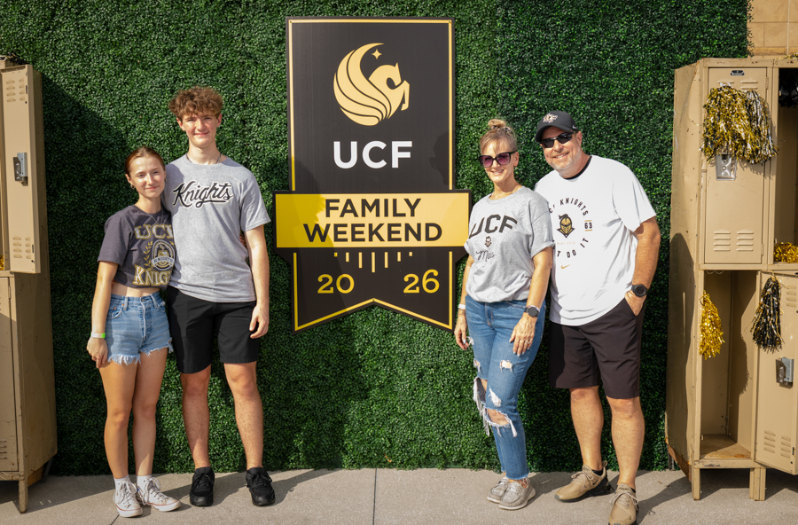 Four people pose in front of a UCF Family Weekend sign on a green hedge backdrop with gold lockers on the sides and gold decorations nearby.