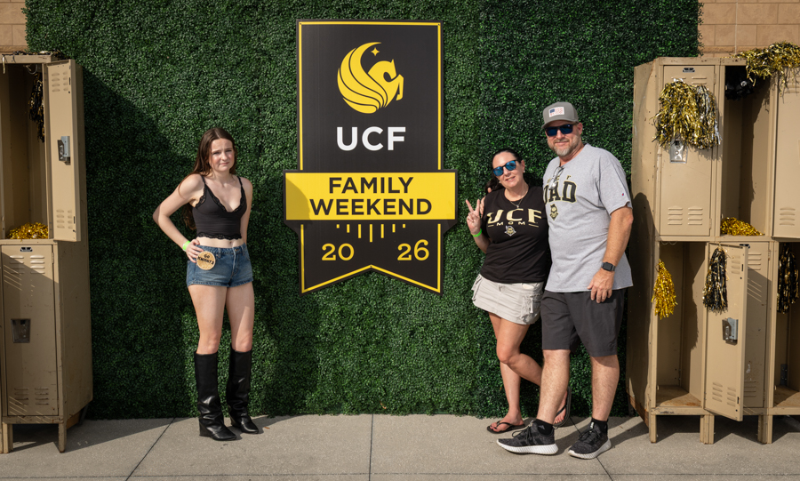 Three people pose in front of a large UCF Family Weekend sign on a green hedge backdrop, year 2026 visible.