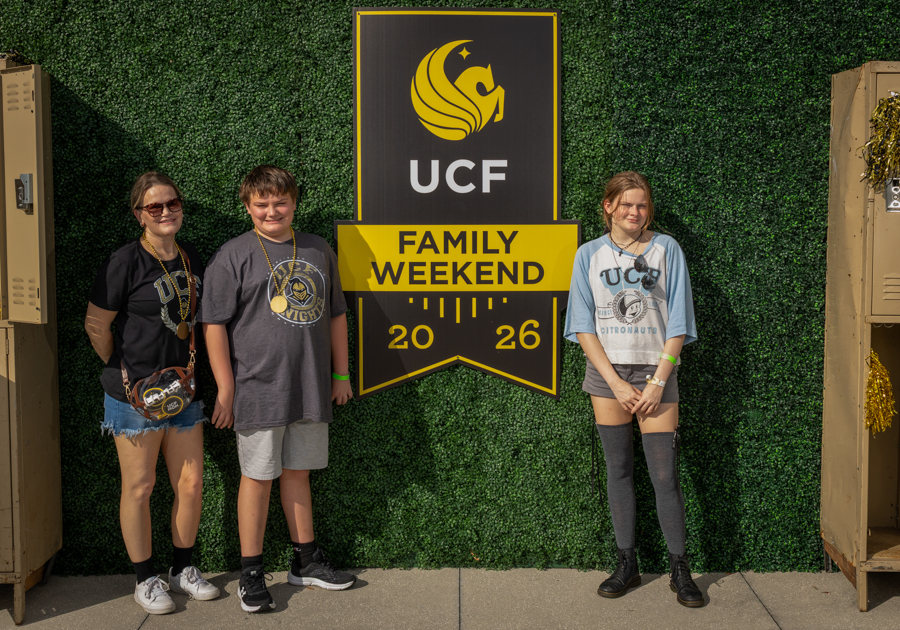 Three people pose beside a yellow and black UCF 'Family Weekend' sign reading 2026 against a green hedge backdrop.