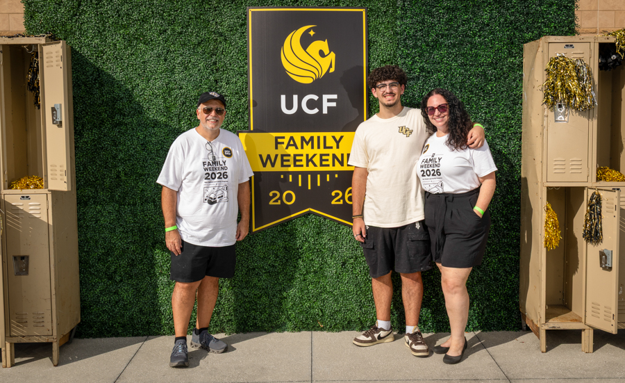 Three people stand in front of a UCF 'Family Weekend 2026' banner: an older man in a white T-shirt and cap on the left, a younger man in a beige T-shirt in the middle, and a woman in a white T-shirt and black shorts on the right, with lockers and greenery backdrop and festive gold decorations.