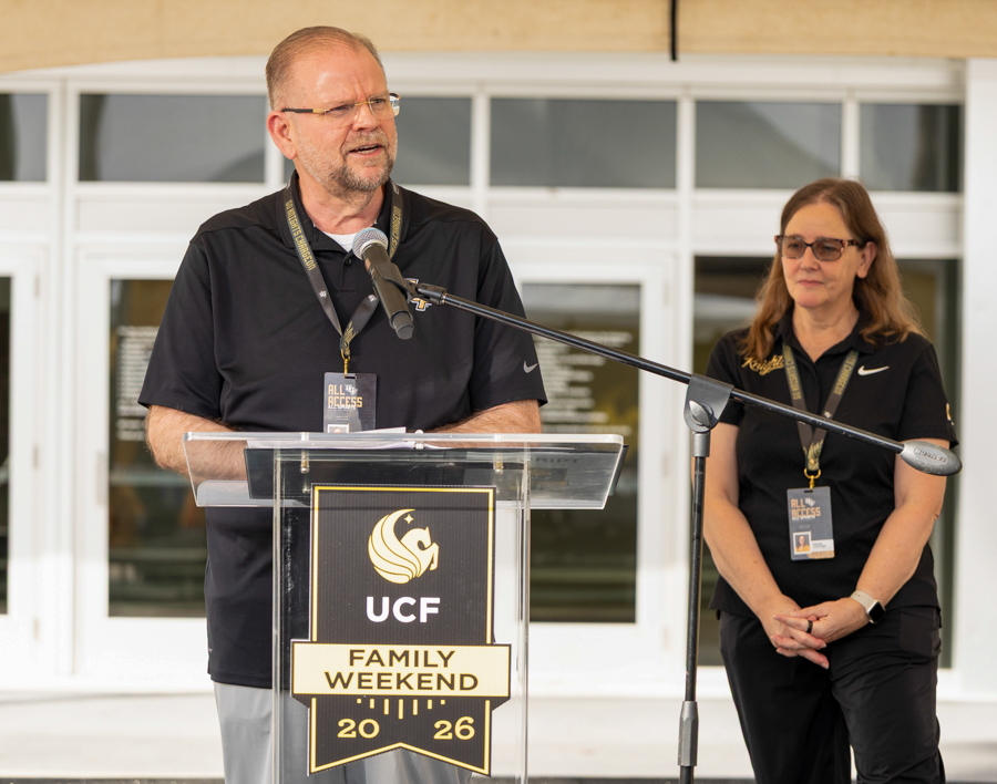 Man speaks at a clear podium with a UCF Family Weekend 2026 banner; a woman stands nearby in a black polo with a lanyard outside the building.