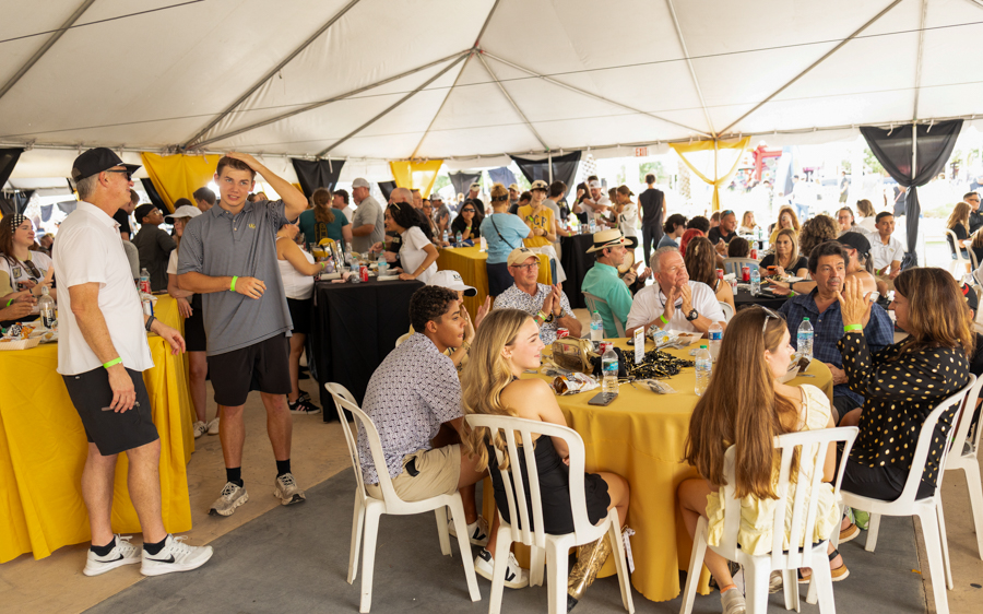 Group of people seated around yellow-clothed round tables at a crowded daytime banquet under a large white tent, socializing and dining.