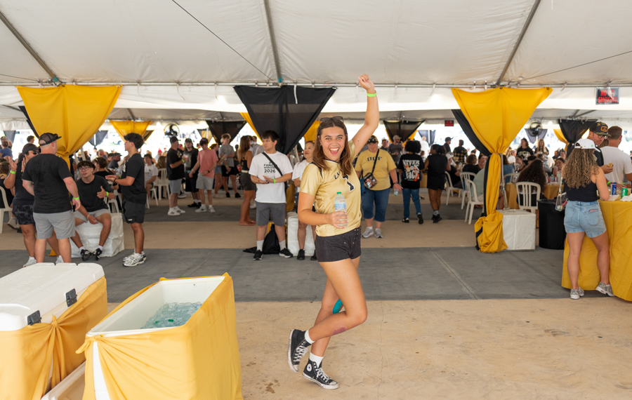 Young woman in yellow shirt and black shorts jumping with a water bottle at a crowded outdoor festival under a white tent.
