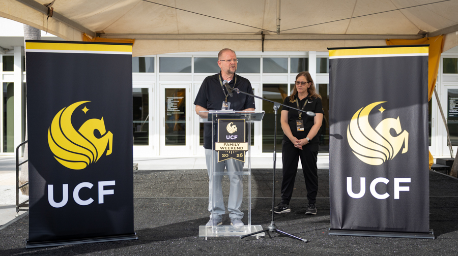 Man speaks at a clear podium with a UCF logo during an outdoor event under a white tent, flanked by large UCF banners and a staffer nearby.