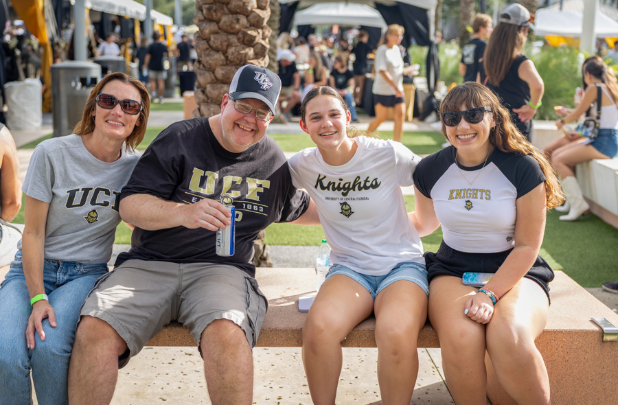 Four people sit on a bench at an outdoor campus event, wearing UCF Knights shirts and cap, smiling at the camera with tents in the background.