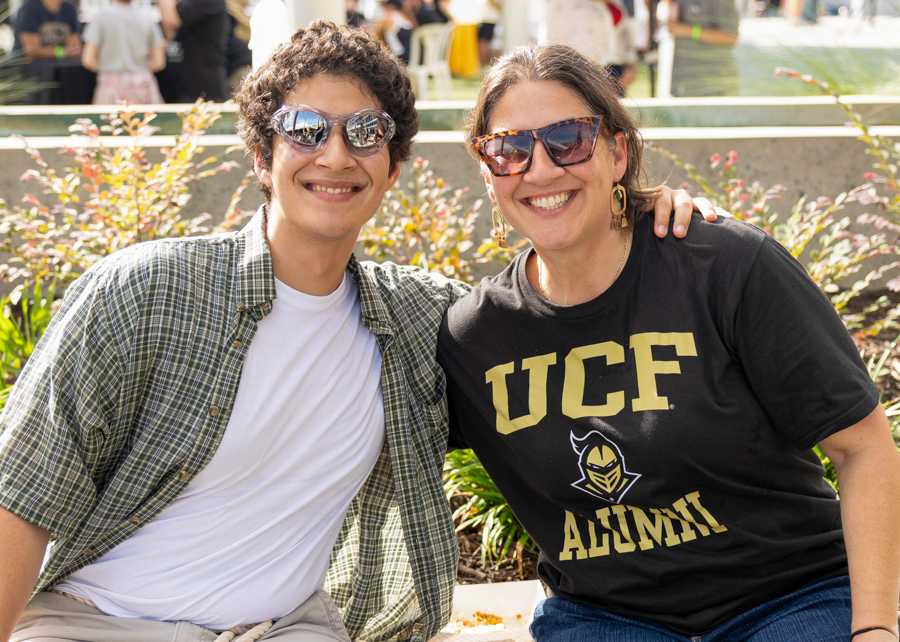 Two friends pose outdoors, smiling at the camera; one in a plaid shirt, the other in a black UCF Alumni shirt with sunglasses, plants in the background.