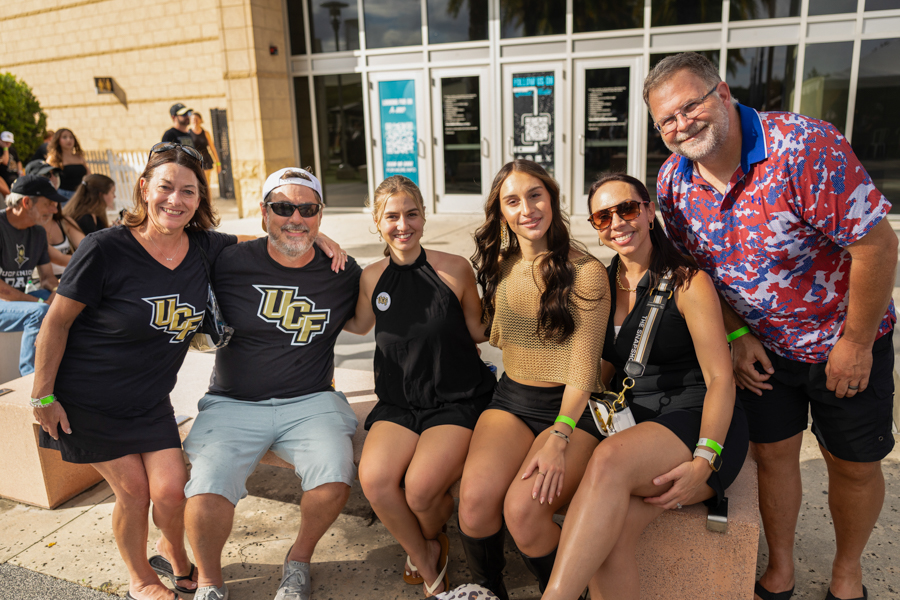 Seven friends posing for a photo outside a campus building, some wearing black UCF shirts, smiling and seated on a low ledge with others standing nearby.