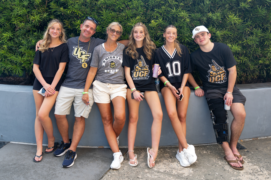 Six friends posing together outdoors in front of a green hedge, wearing UCF Knights apparel and smiling for the photo.