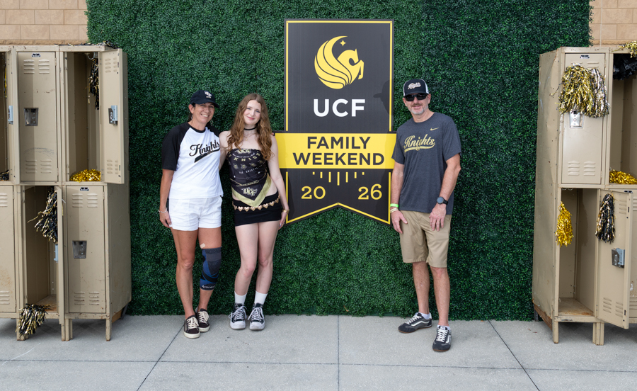 Three people pose in front of a UCF Family Weekend sign against a green hedge wall, with gold lockers on either side.