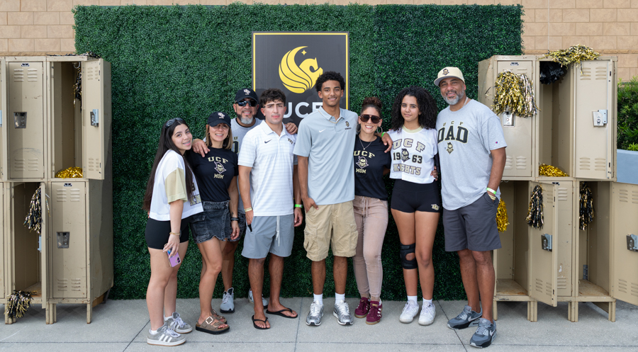 Group portrait of ten people posing together in front of a green hedge wall with a crest sign, flanked by lockers decorated with gold tinsel and bows on both sides, outdoors.