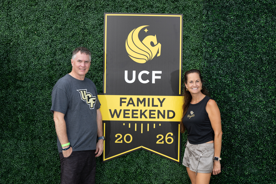 Two adults stand on either side of a large UCF Family Weekend 2026 sign against a green hedge backdrop.