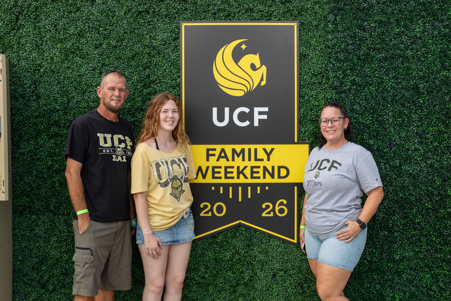 Three people pose in front of a UCF Family Weekend 2026 sign against a green hedge wall.