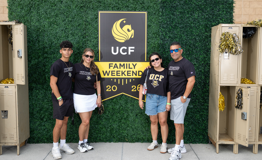 Family of four posing in front of a UCF Family Weekend banner with a green hedge backdrop and lockers on the sides.