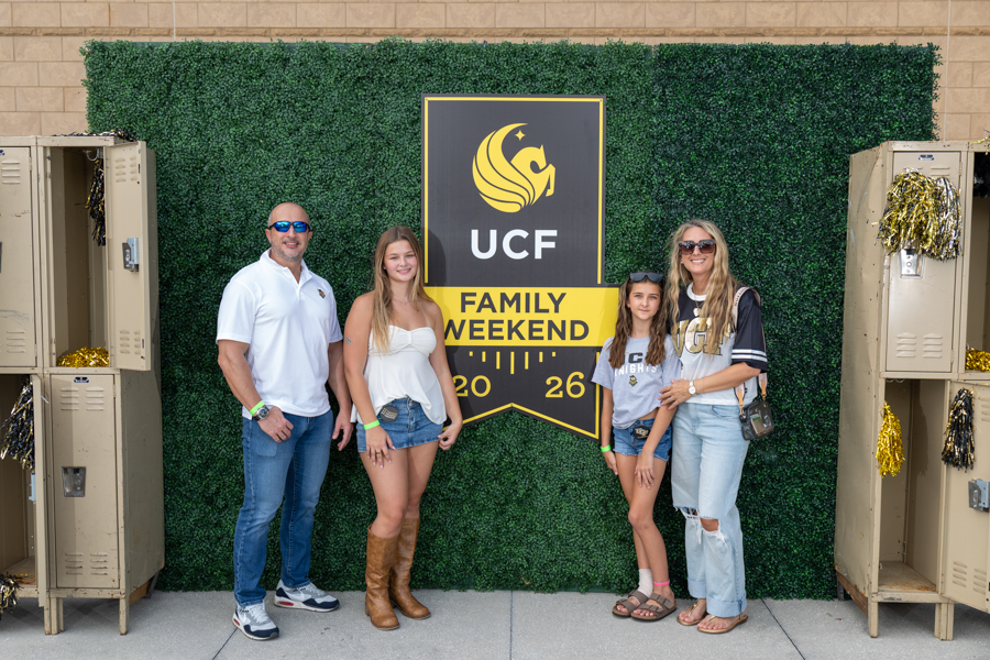 Family of four posing in front of a UCF Family Weekend banner on a green hedge backdrop, with lockers on both sides.