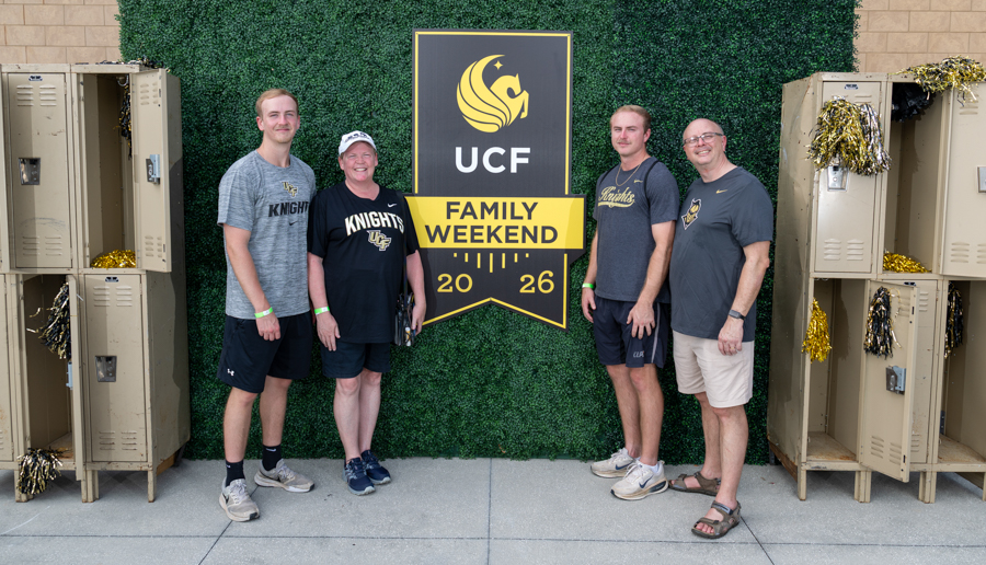 Five adults pose for a photo in front of a UCF Family Weekend banner, wearing Knights apparel and casual outfits.
