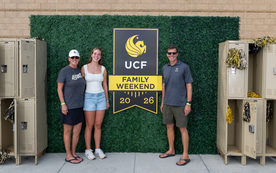 Three people pose beside a large UCF Family Weekend sign on a green hedge backdrop, with beige lockers on both sides.