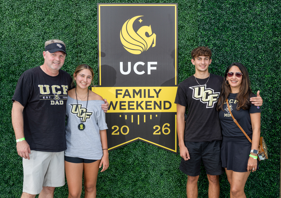 Family of four posing in front of a UCF Family Weekend banner on a green hedge backdrop, all wearing UCF apparel (2026).