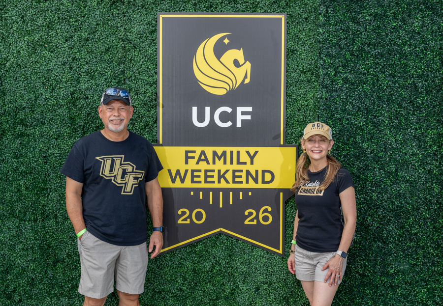 Two smiling adults stand beside a large UCF Family Weekend sign on a green hedge backdrop, 2026 calendar shows 20 and 26.
