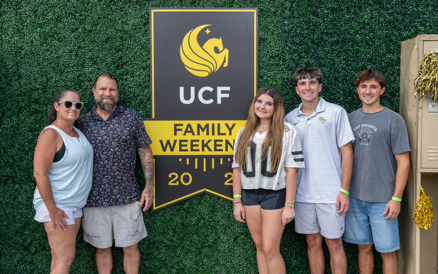 Five people pose together in front of a UCF Family Weekend banner on a green hedge background, 2020 inset.