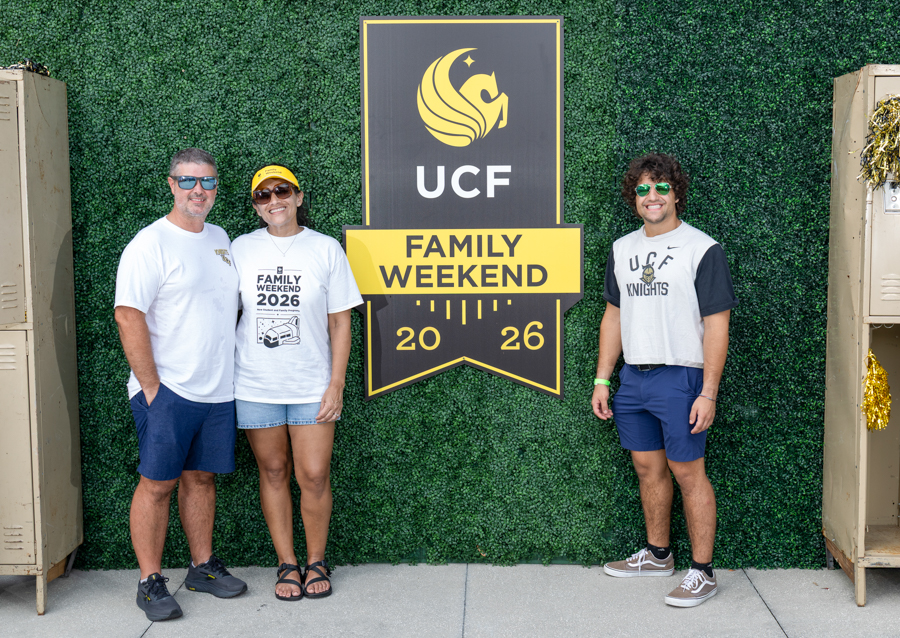 Three people pose beside a large UCF Family Weekend 2026 sign on a green hedge wall, smiling at the camera (two adults on left, one student on right).