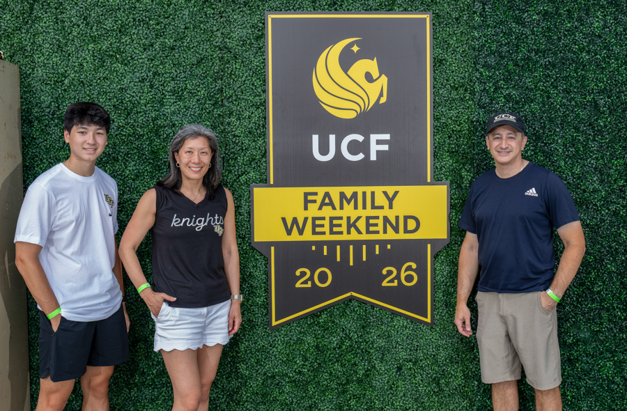 Three people stand beside a large UCF Family Weekend sign mounted on a green hedge backdrop, posing for a photo together.